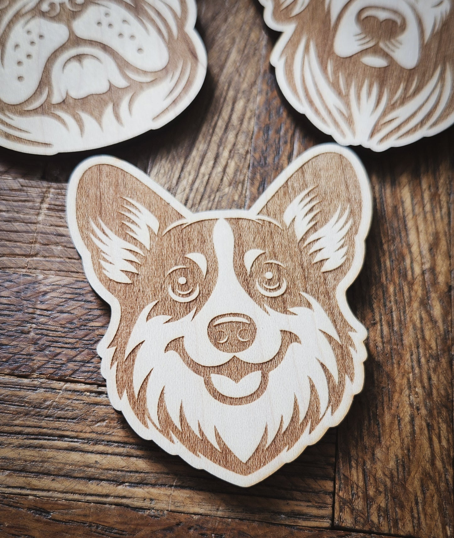 An engraved magnet of a corgi on a wooden background, with engraved magnets of a golden retriever and an English bulldog in the background